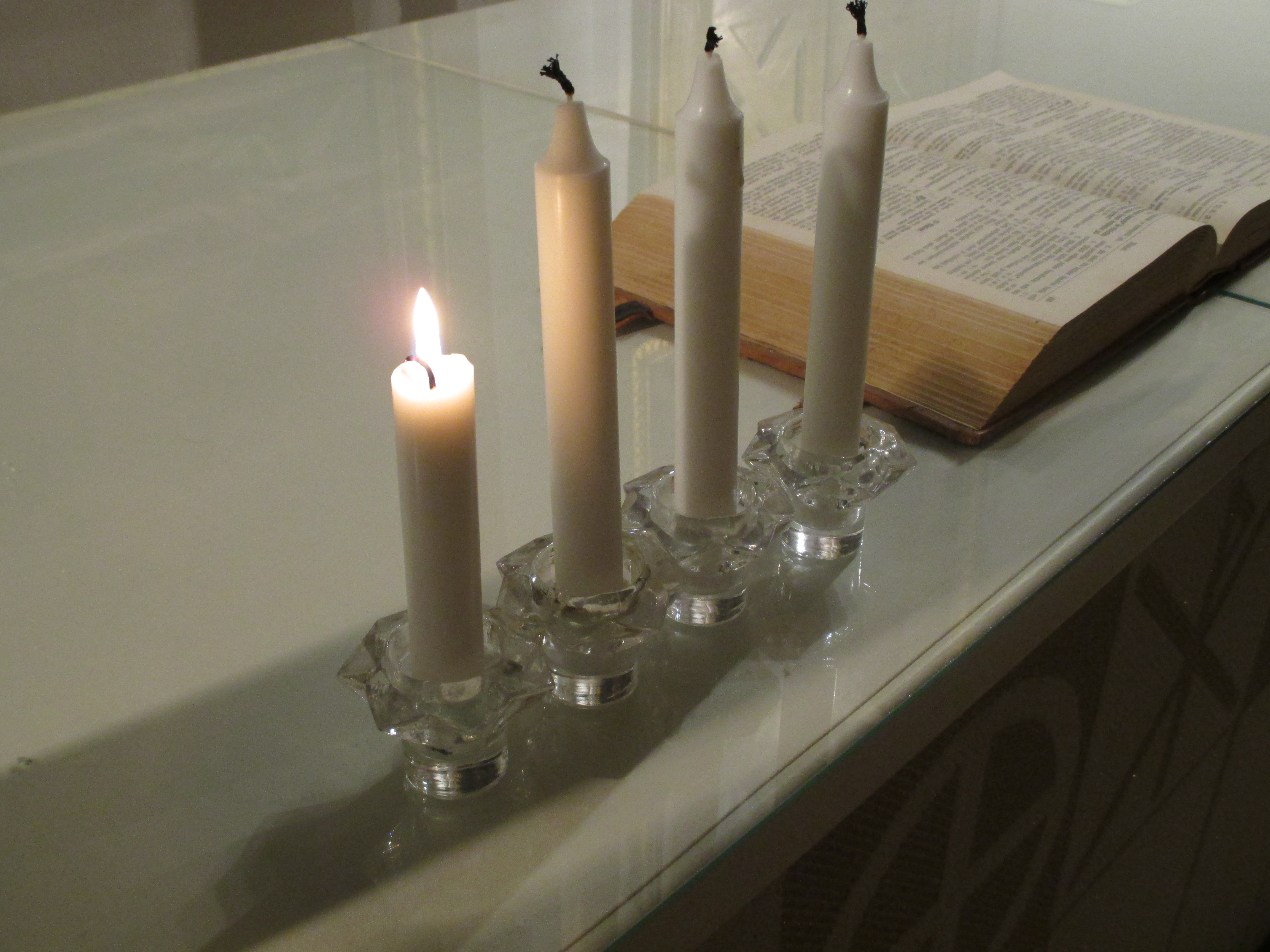 Valkoisella liinalla katetulla pöydällä on lasisissa kynttilänjaloissa rivissä neljä kynttilää ja taempana avattu Raamattu.  Sytytettynä on vain yksi kynttilä, joka on ehtinyt palaa lyhyemmäksi kuin muut.