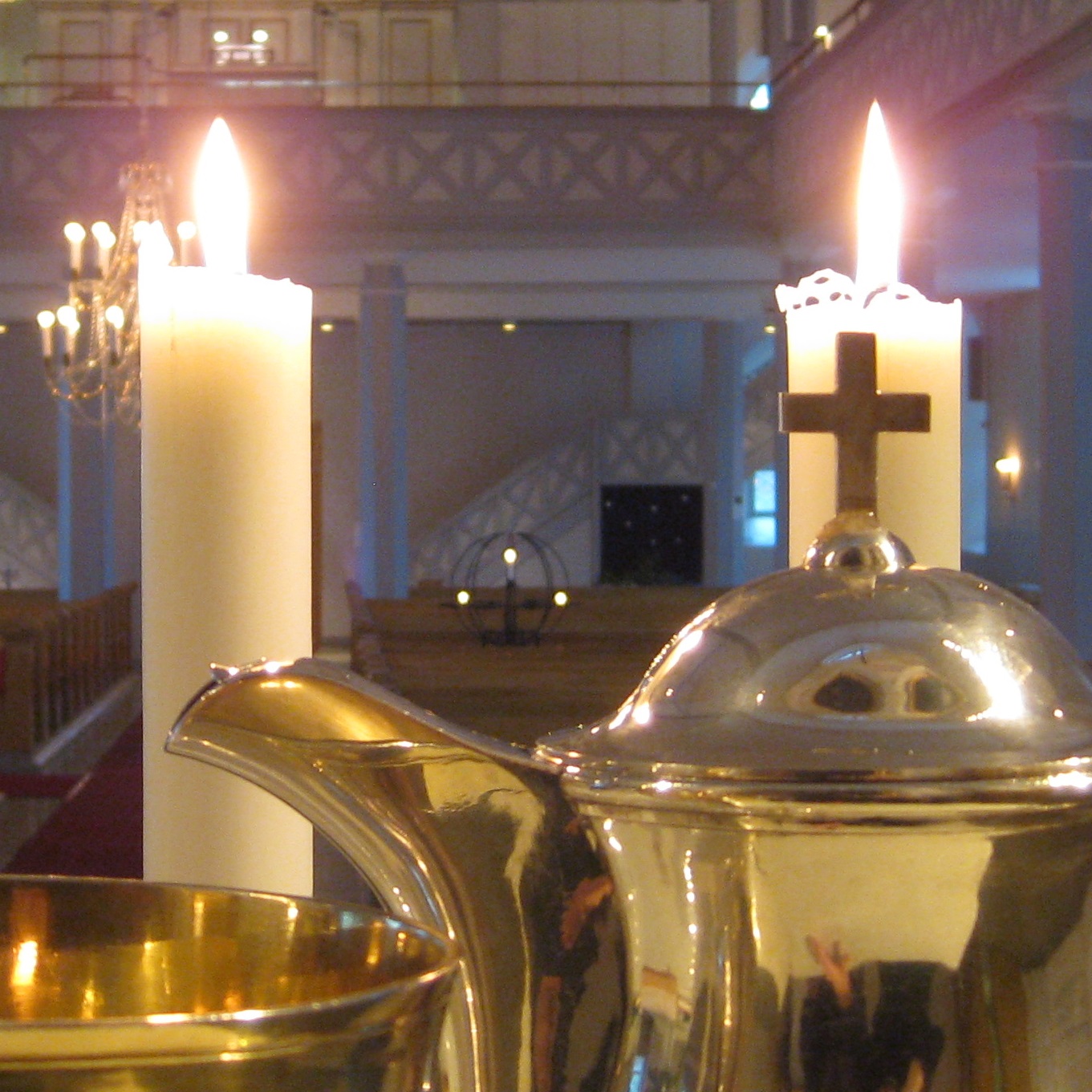 etiualalla ehtoollisviinikannu ja kaksi palavaa kynttilää, taempana kirkonpenkkejä ja urkuparvi