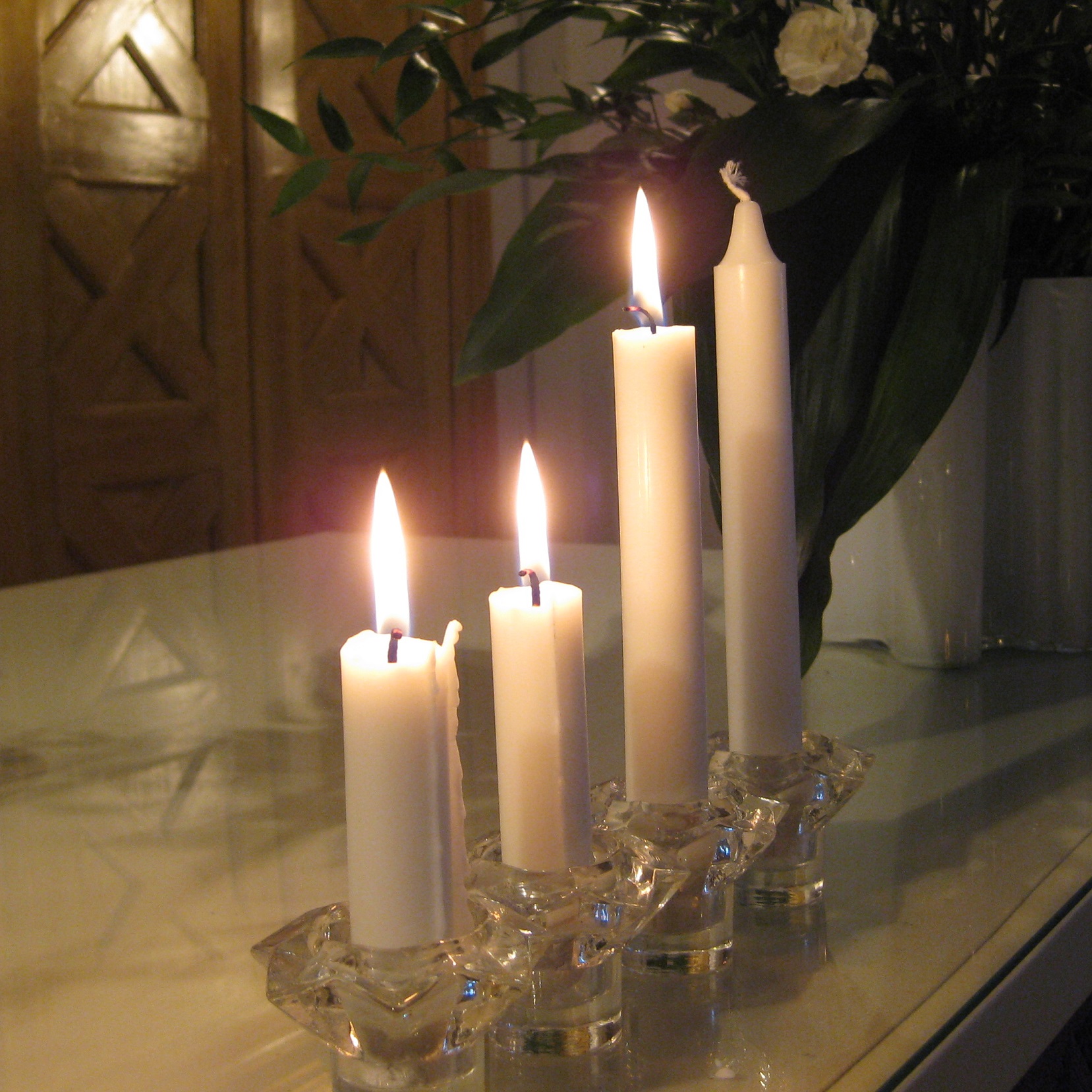 Neljä valkoista kynttilää on rivissä pöydällä, kolmessa liekki ja yksi vielä sytyttämättä.  Palavista kynttilöistä kaksi on jo melko lyhyitä.