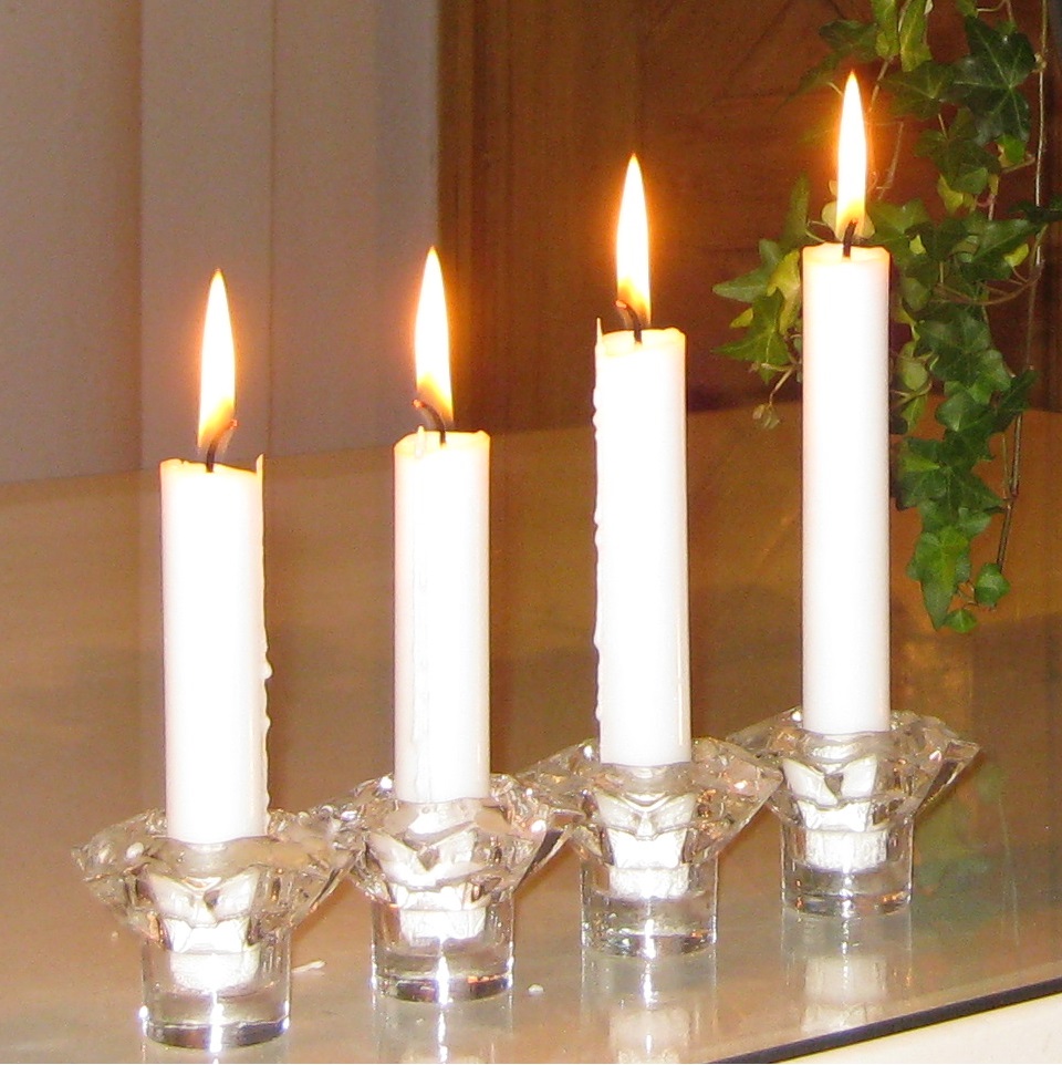 Neljä valkoista, eripituista kynttilää on pöydällä rivissä palamassa, oikealla sivulla viherkasvin rönsyjä.