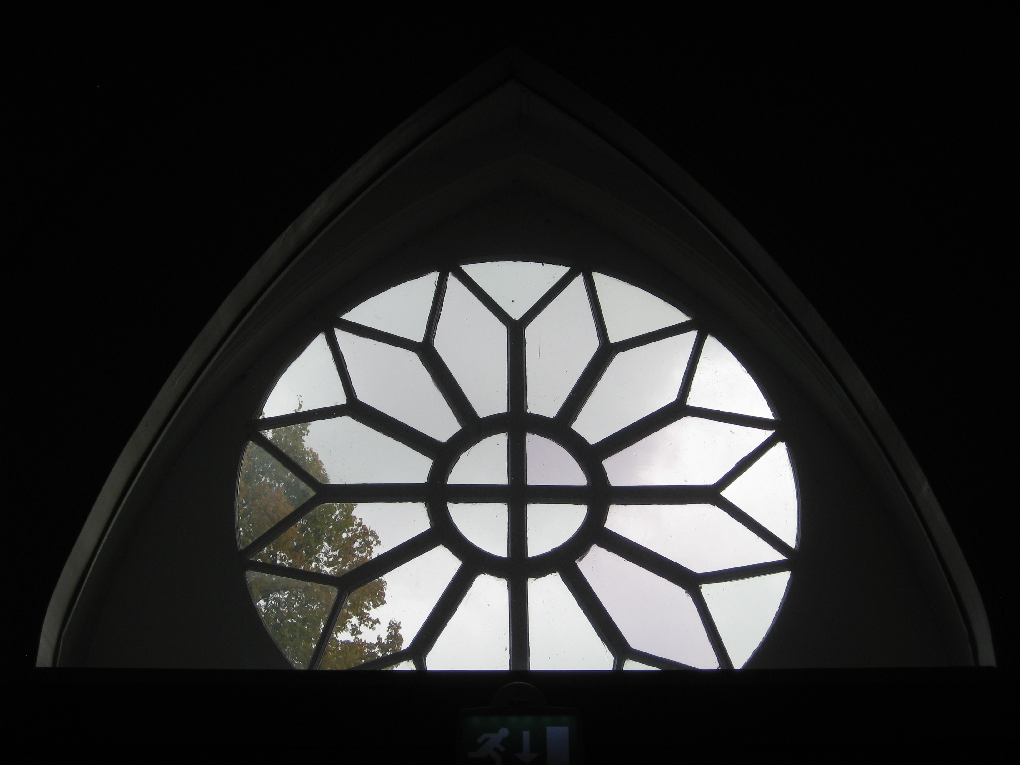 Pyöreä ikkuna, jonka keskellä on ristikuvio.  Kuva on otettu hämärästä sisätilasta; ikkunasta näkyy päivänvaloa ja puun latvaa.