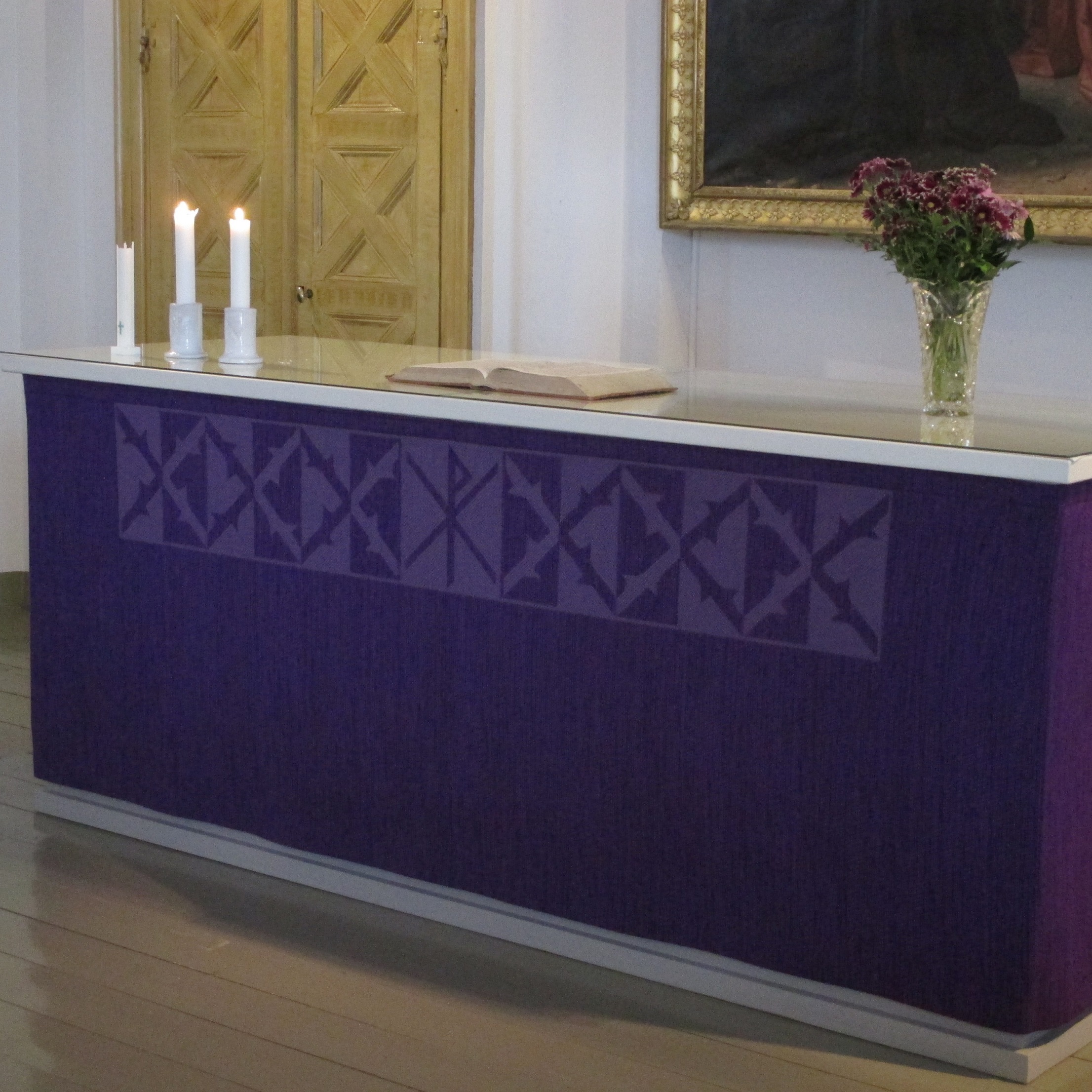 violettiin kankaaseen verhoiltu alttaripöytä, jolla on kukkia, avoin Raamattu ja kaksi palavaa kynttilää
