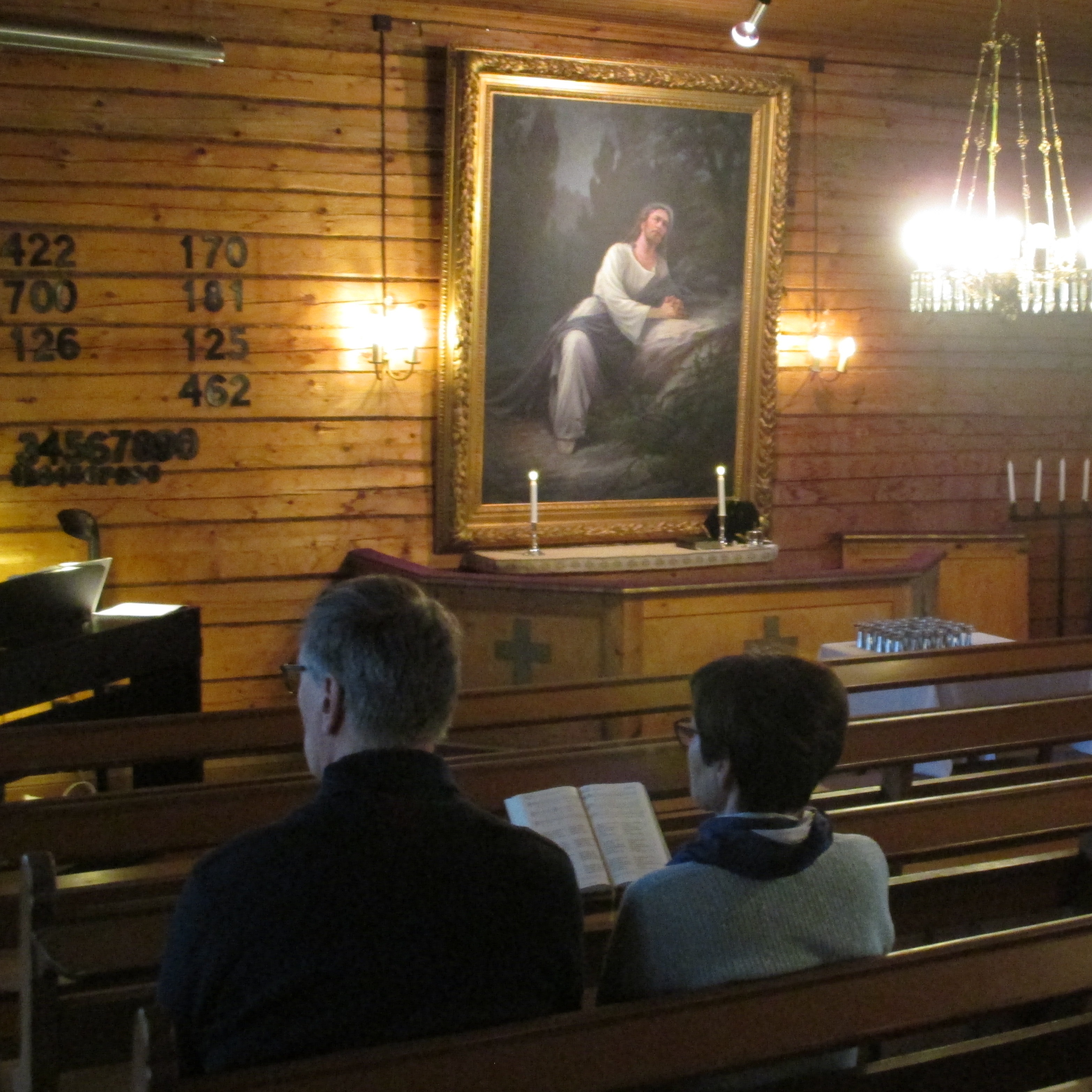 Pariskunta istuu kirkonpenkissä.  Heidän edessään hirsiseinälle on ripustettu virsinumeroita sekä rukoilevaa Jeesusta esittävä alttaritaulu