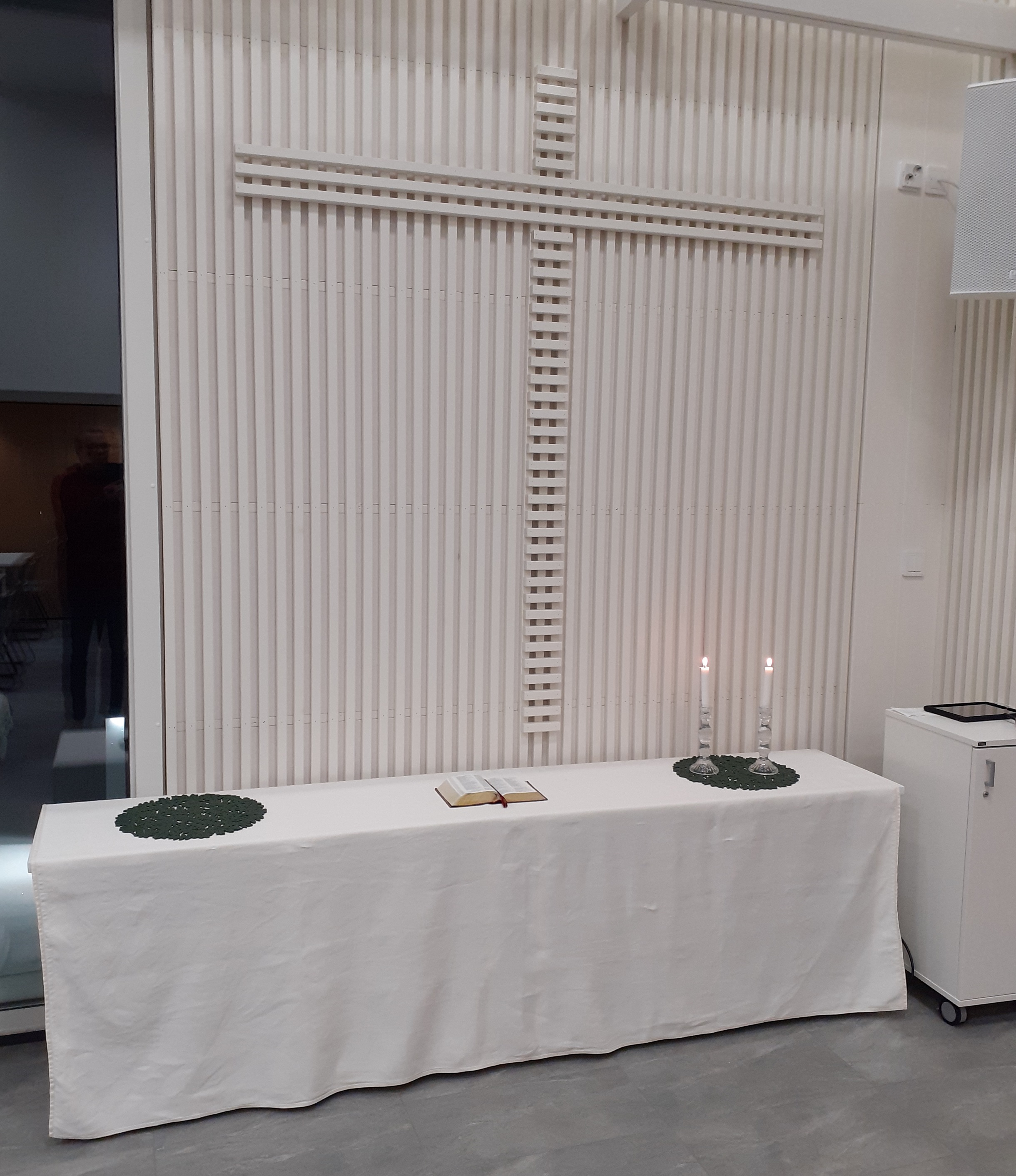 vaalea seinä, jossa on iso ristikuvio ja sen alla seinää vasten pöytä, jolla avattu Raamattu ja kaksi palavaa kynttilää
